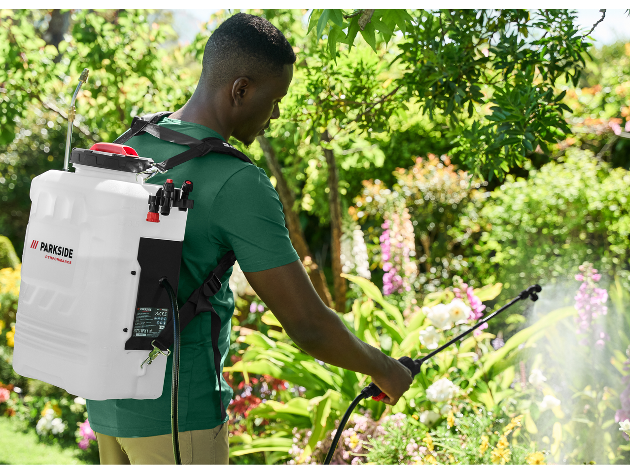 Man in a garden using a backpack sprayer to water plants, with a visible logo on the sprayer.