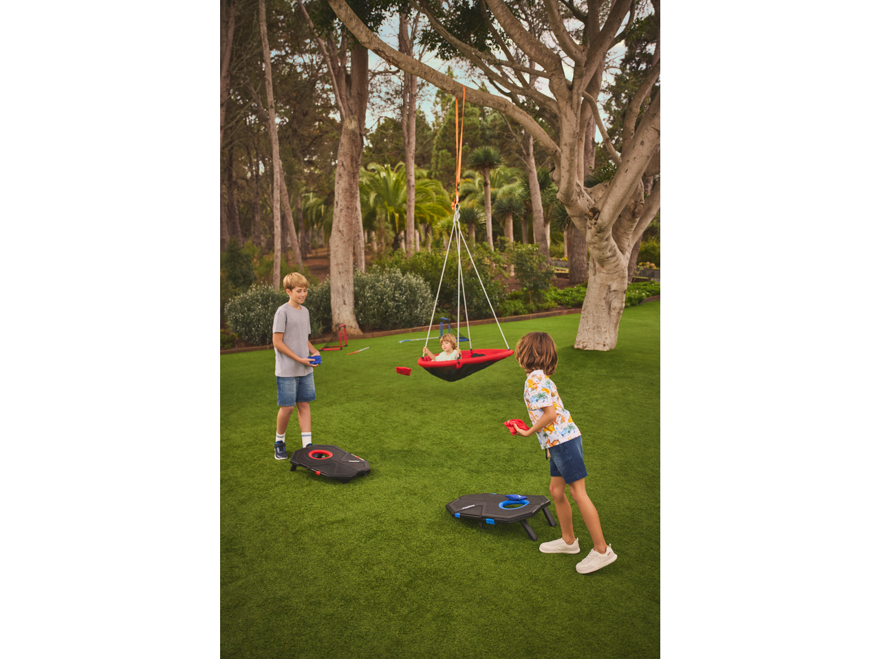 Kids playing bean bag toss on artificial grass, with one child in a swing.