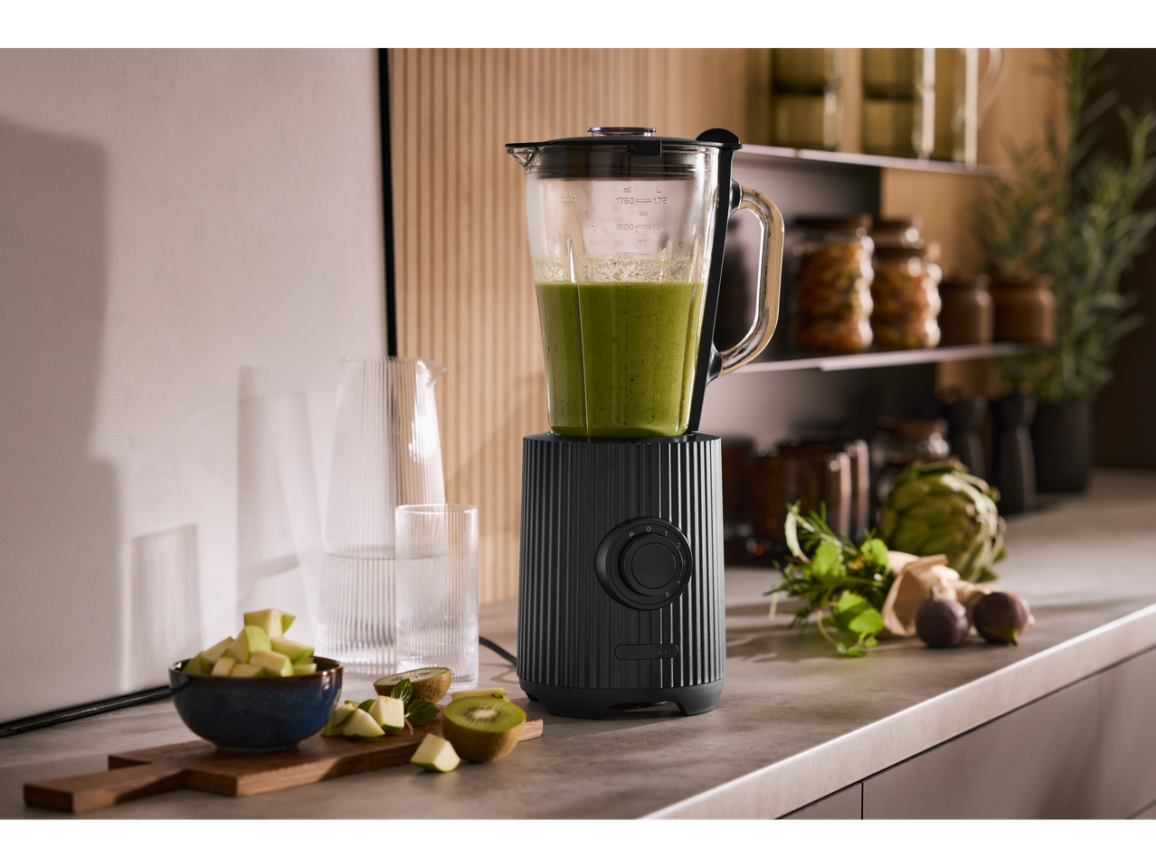 Blender cu smoothie verde, fructe și legume pe un blat de bucătărie.