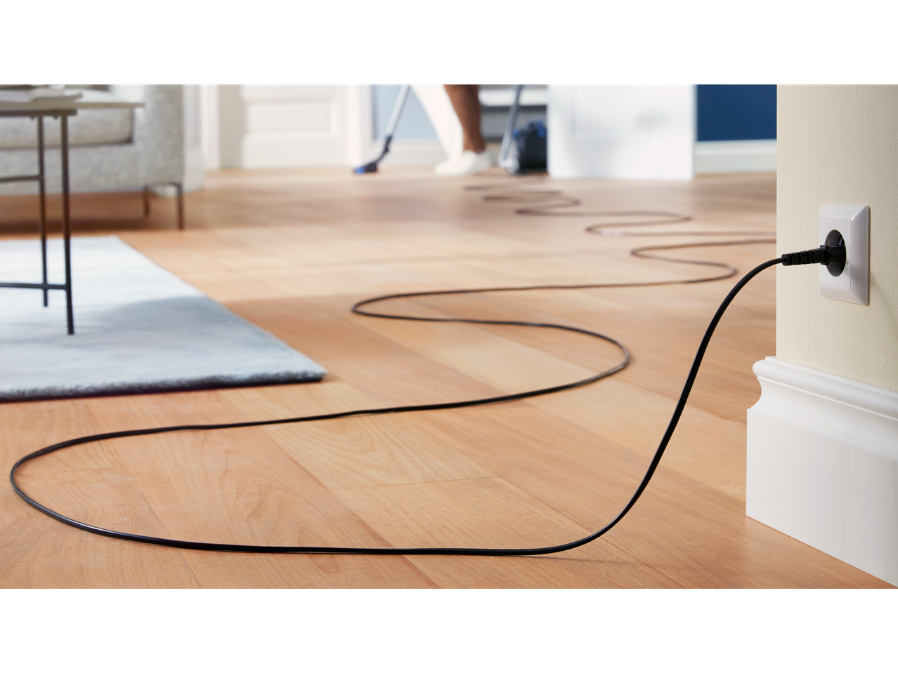 Een zwarte stroomkabel loopt over een houten vloer naar een stopcontact, terwijl op de achtergrond iemand aan het stofzuigen is.