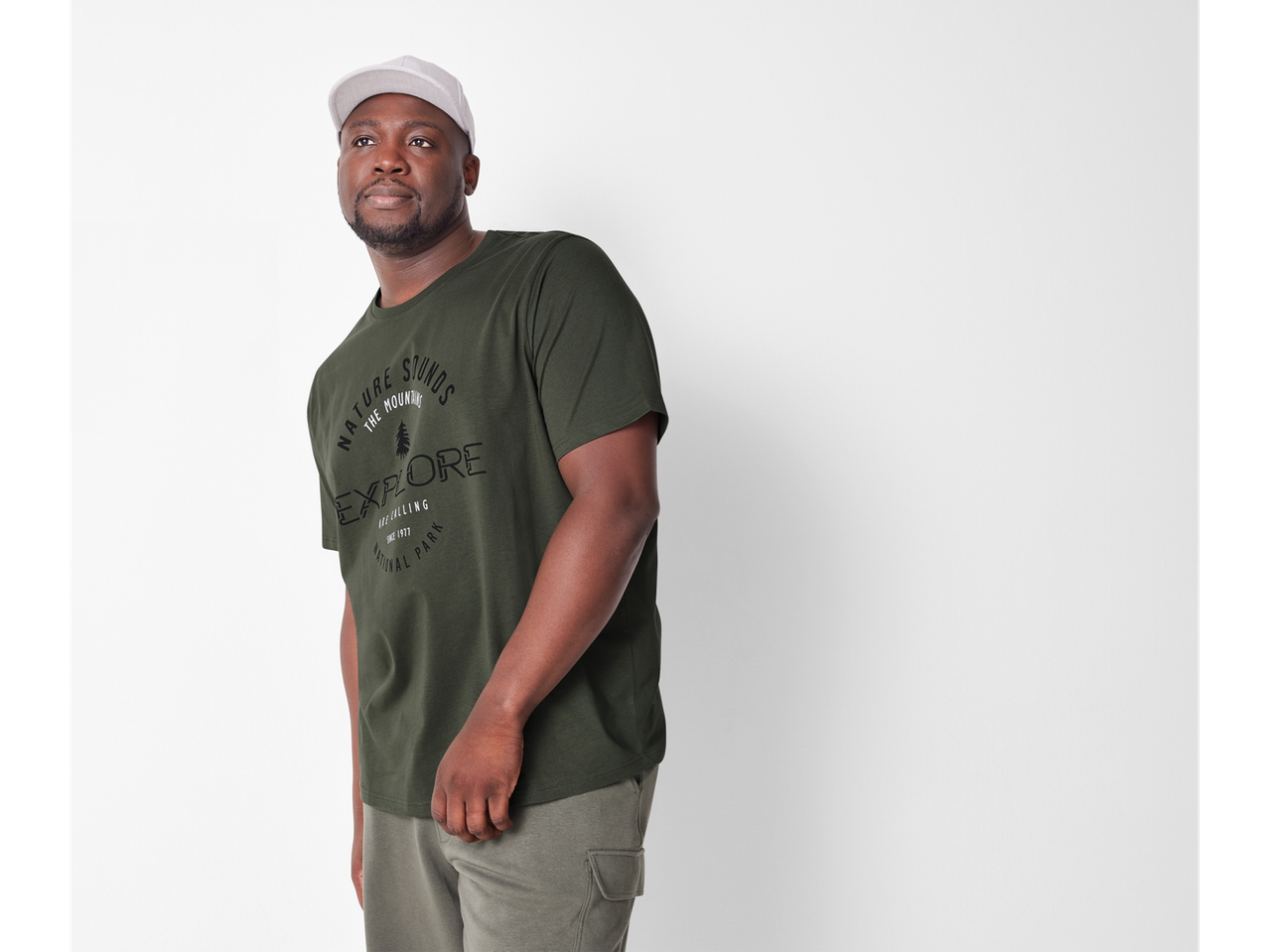 Un bărbat poartă un tricou verde-oliv cu imprimeu și o șapcă de baseball.