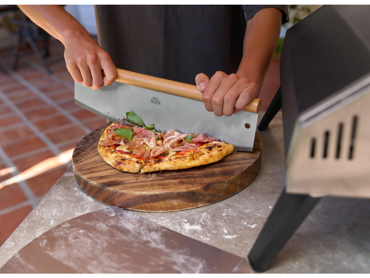 Grillmeister pizza cutter slicing a pizza with prosciutto and basil on a wooden board.