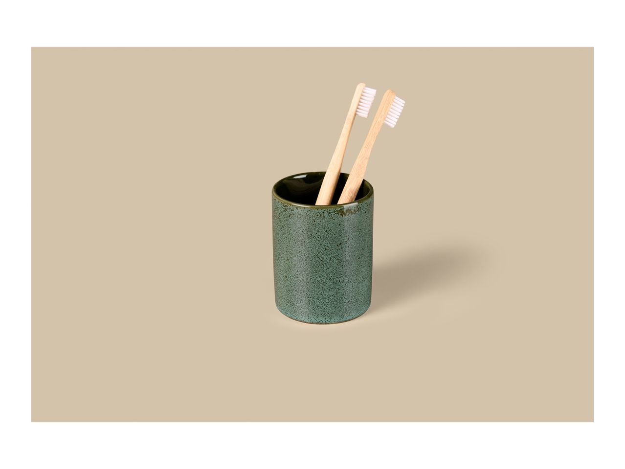 Two bamboo toothbrushes in a green ceramic holder on a beige background.