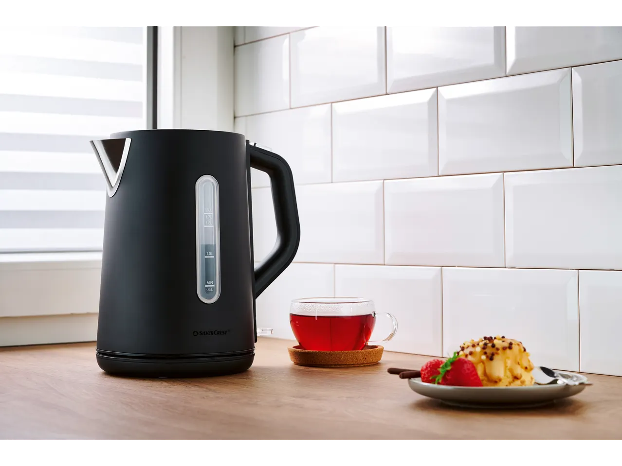 Black SILVERCREST® electric kettle with a cup of tea and a cake on a wooden countertop.