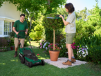 Мъж коси трева, докато жена подрязва храст с електрически ножици в градина.