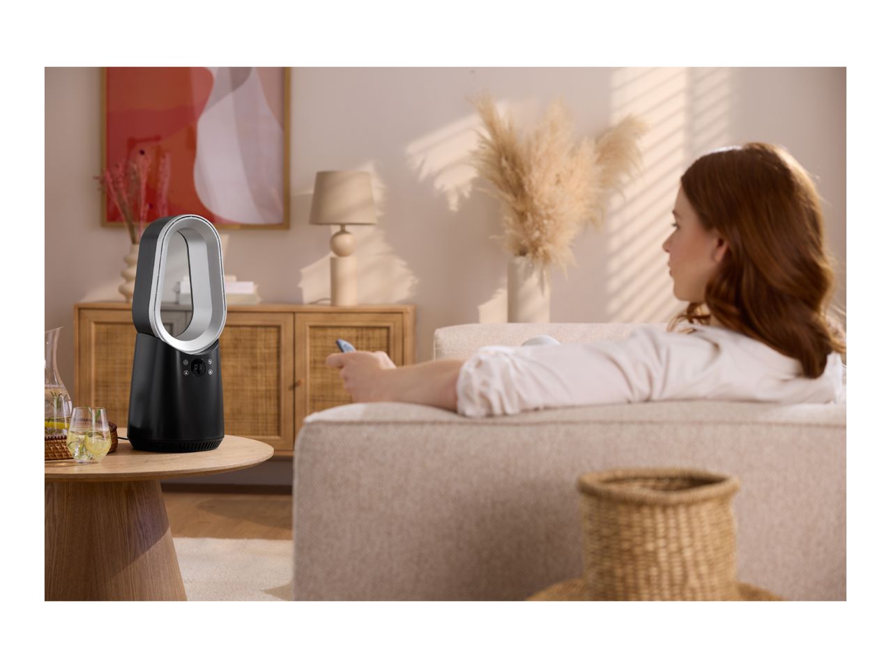 A woman relaxing on a sofa, using a remote control for a black and white air purifier on a side table.