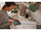 A girl uses a stylus on a pink LCD writing tablet while wearing headphones and studying at her desk.