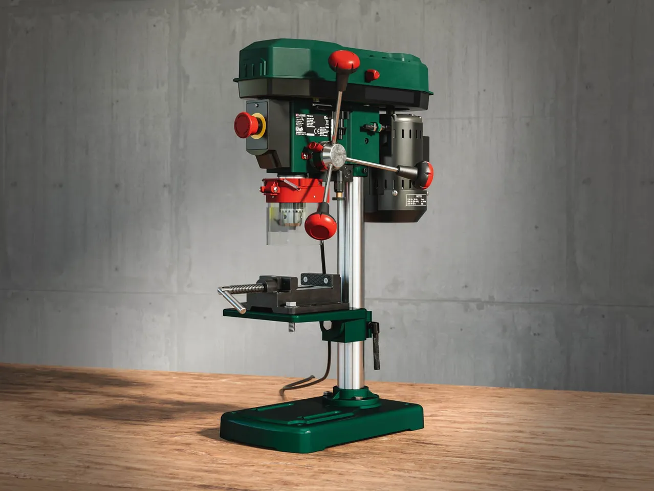 A green and red drill press with a vise on a wooden table against a concrete wall.