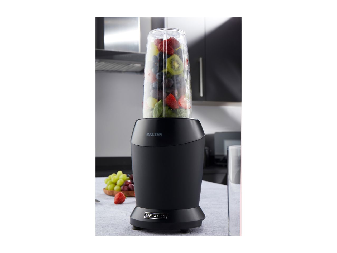 Black blender with a clear cup filled with various fruits, 1000 Watts, in a kitchen.