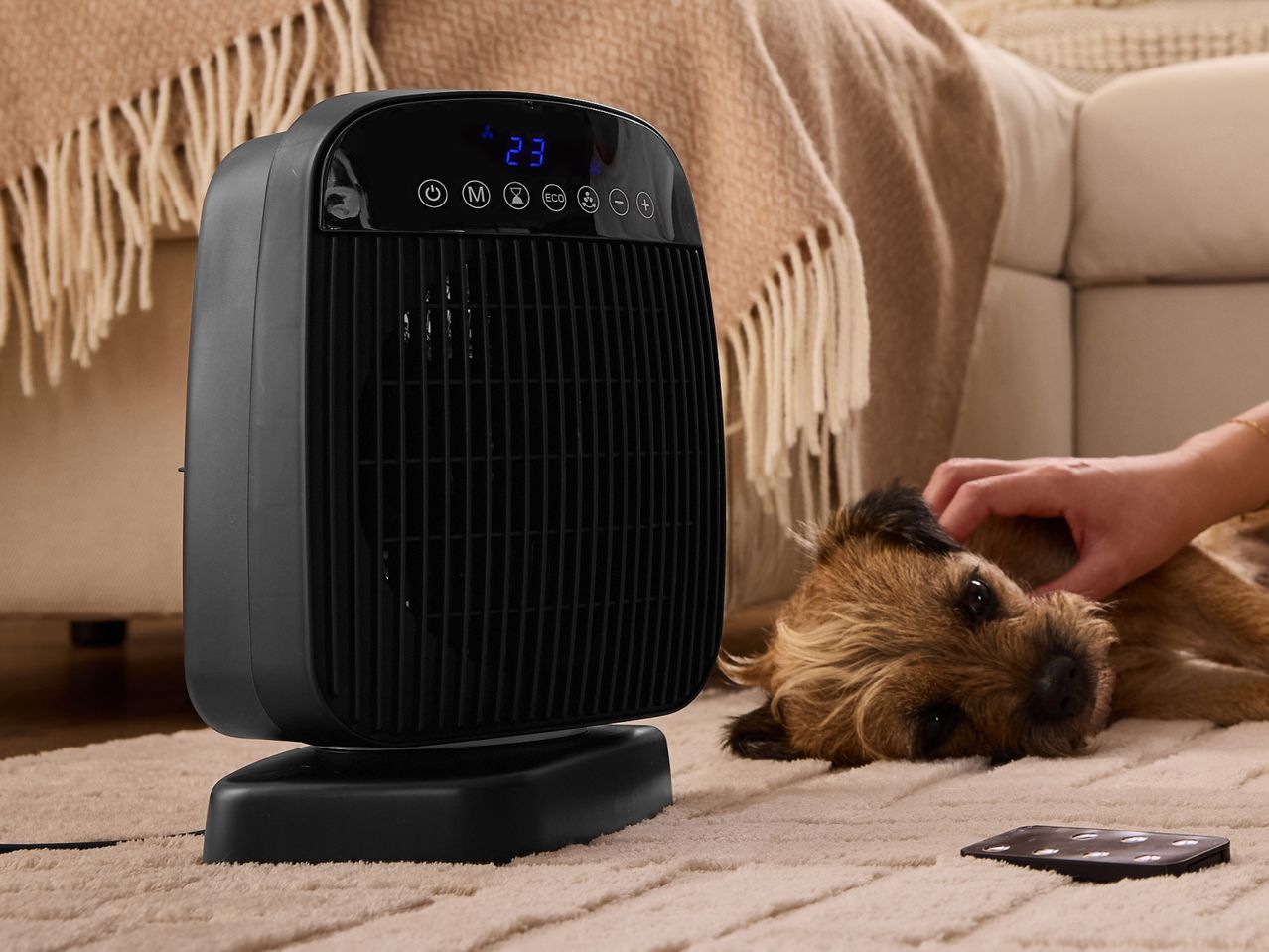Black electric heater with digital display, dog being petted, and remote control on a rug.