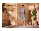 Man in a grey short-sleeved shirt, denim shorts, and sandals, standing in a sunny, desert-themed setting.