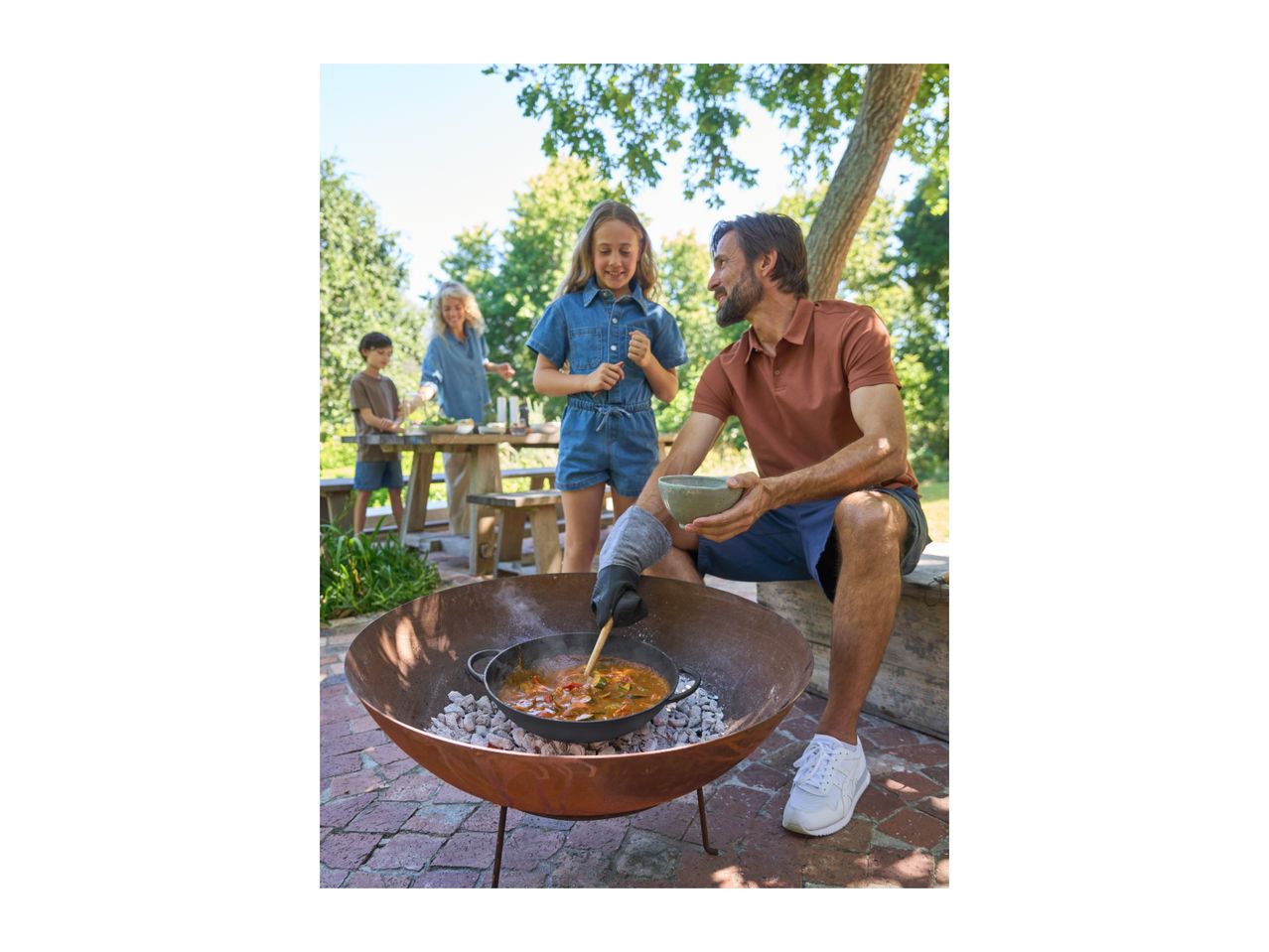 Family cooking outdoors with a fire pit, preparing a meal in a cast iron pot.