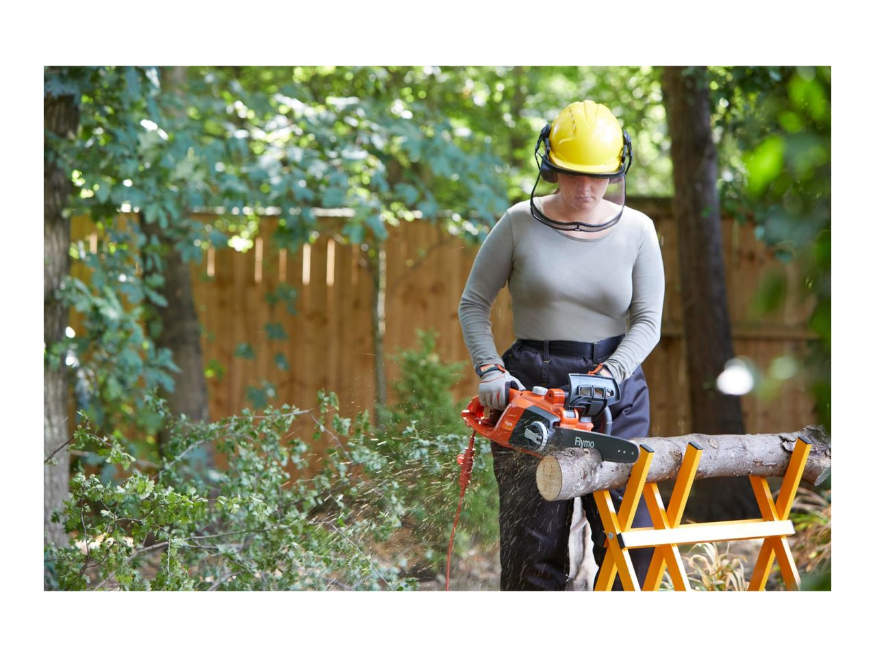 Woman in safety gear cutting wood with a chainsaw on a sawhorse.