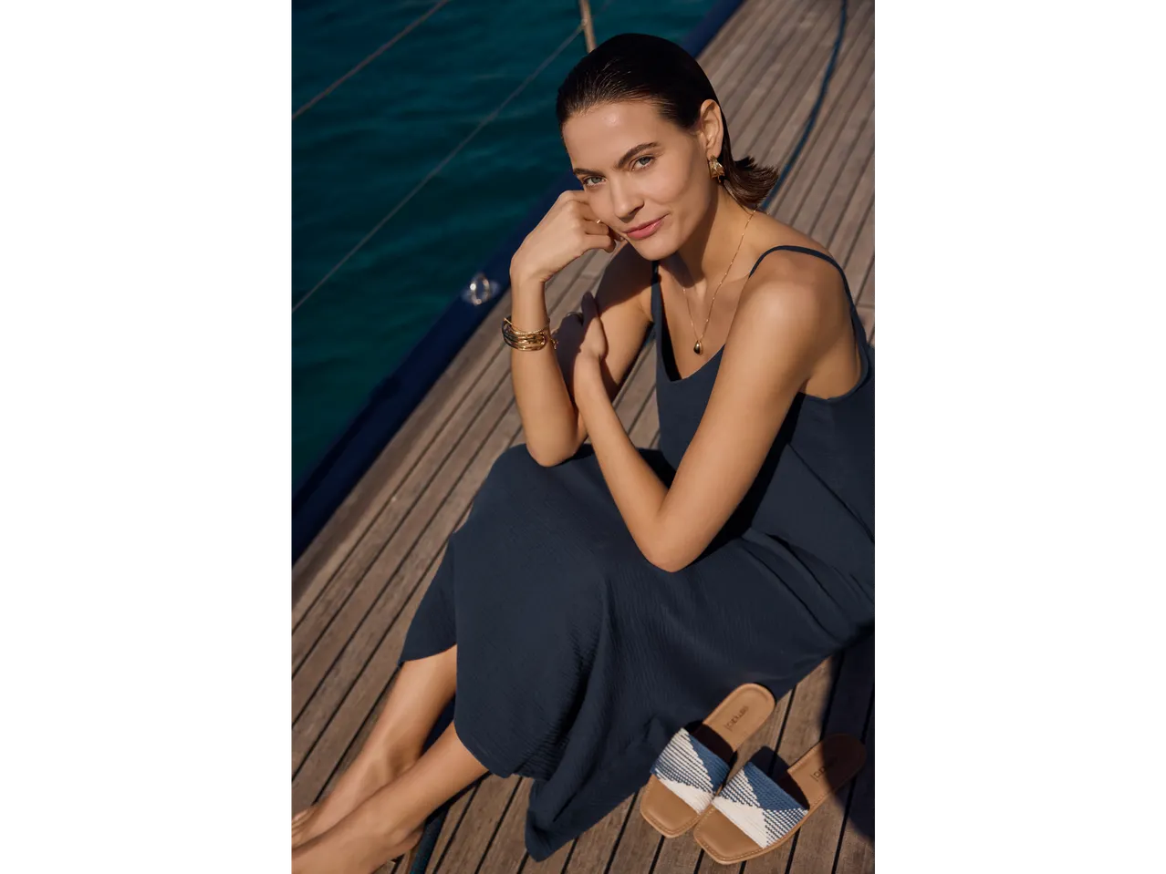 Mulher com vestido azul marinho e joias douradas sentada no convés de um iate usando sandálias Loq.