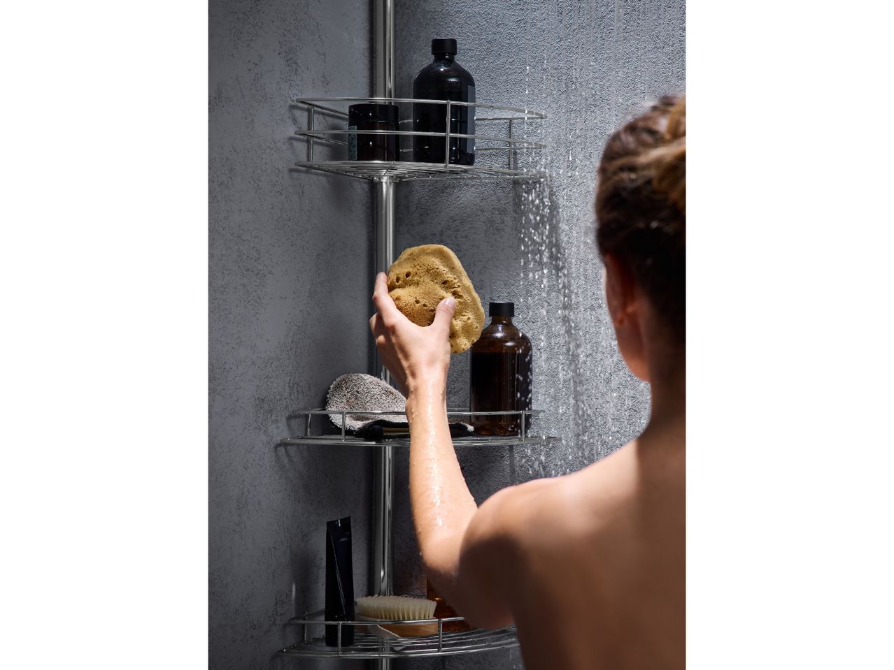 A person showering, holding a natural sponge, with shower caddy and bottles.