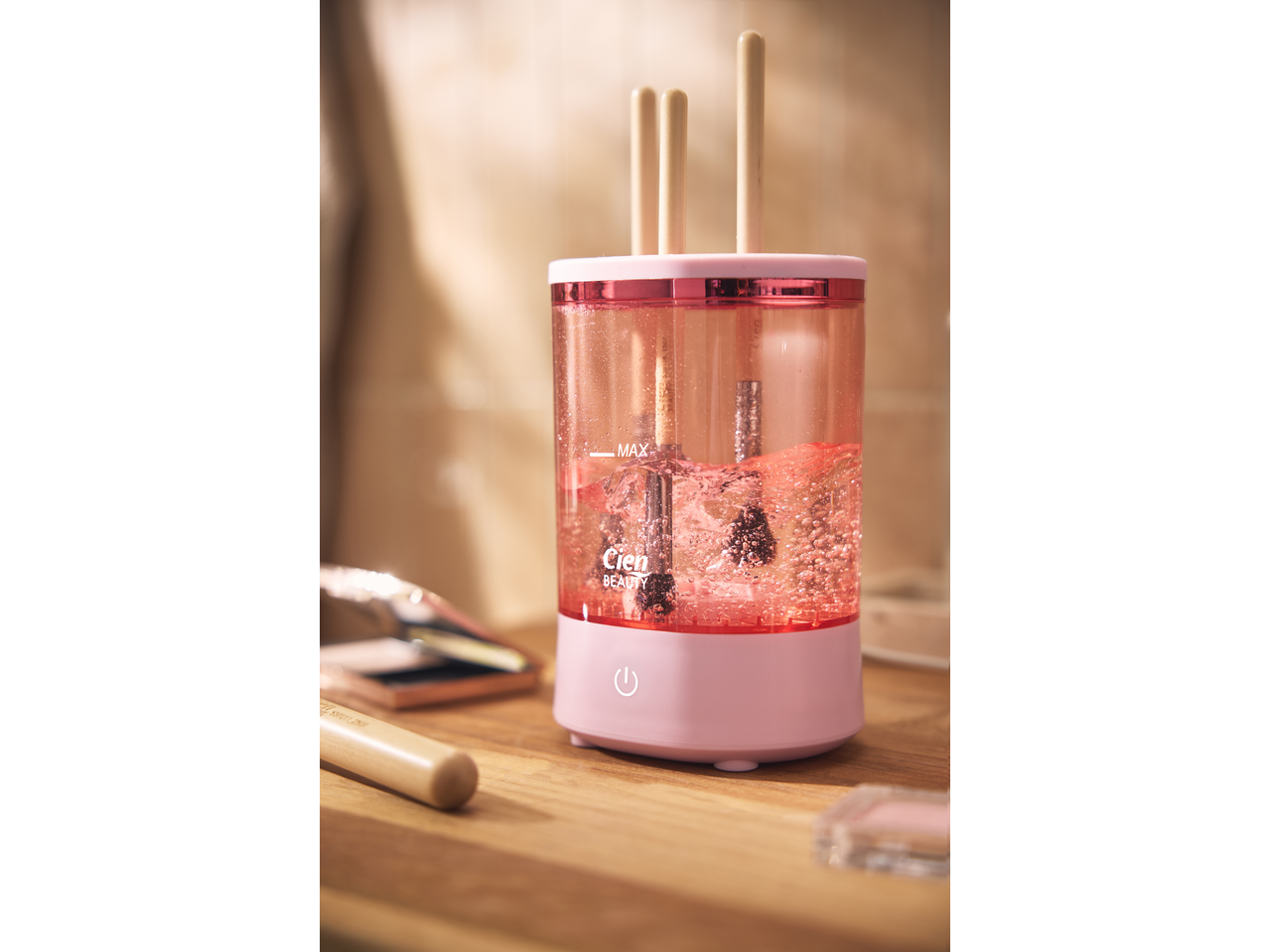 Pink makeup brush cleaner with brushes inside, filled with bubbly pink liquid, on a wooden surface.