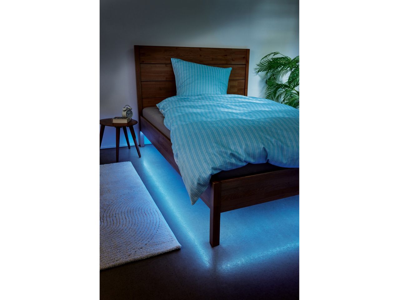 Letto in legno con illuminazione a LED blu sotto, comodino e pianta.