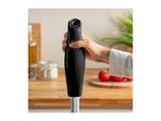 Hand holding a black immersion blender in a kitchen with tomatoes and spices.