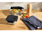 Stainless steel food containers with caprese salad, a wooden pepper mill, and a kitchen towel.