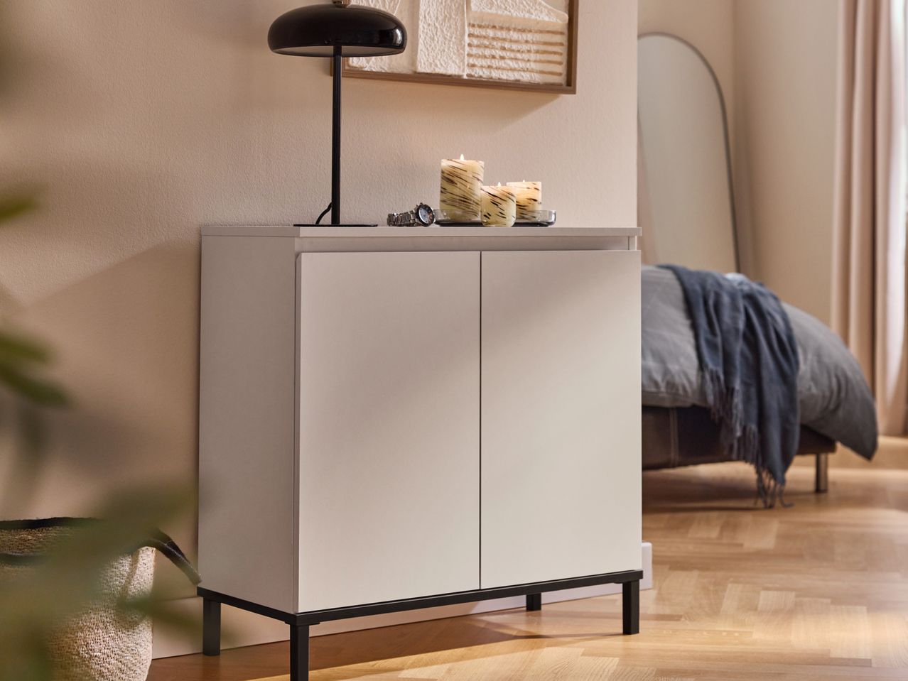 White cabinet with black legs, a lamp, and candles on top, in a bedroom setting.