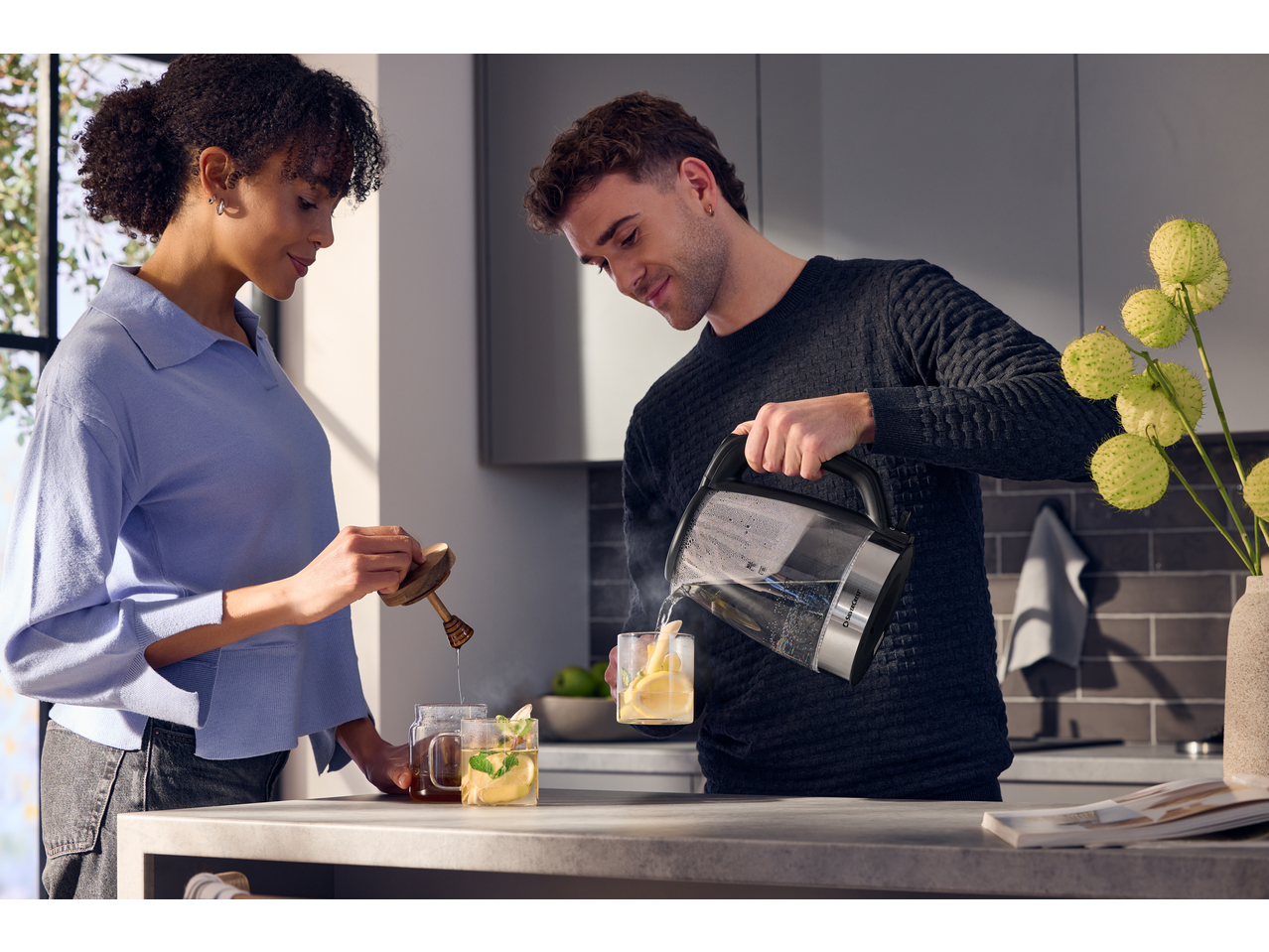 Man pouring water from a SILVERCREST® Colour Changing Glass Kettle into a mug, woman adding honey.