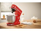 Robot da cucina rosso con ciotola in acciaio inox e ingredienti per la cottura su un tavolo di legno.