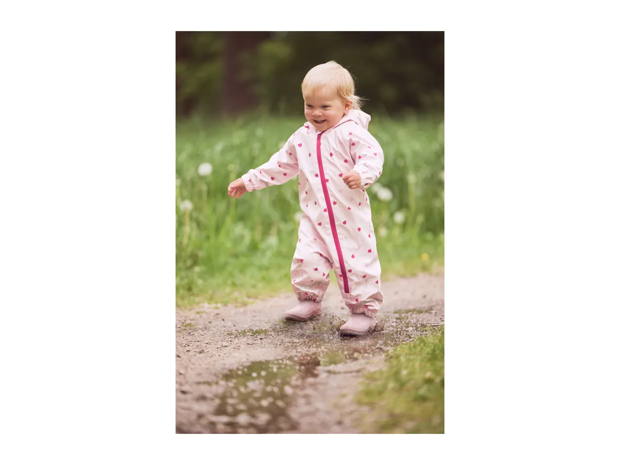 Toddler in a pink heart-patterned waterproof suit and boots splashing in a puddle.