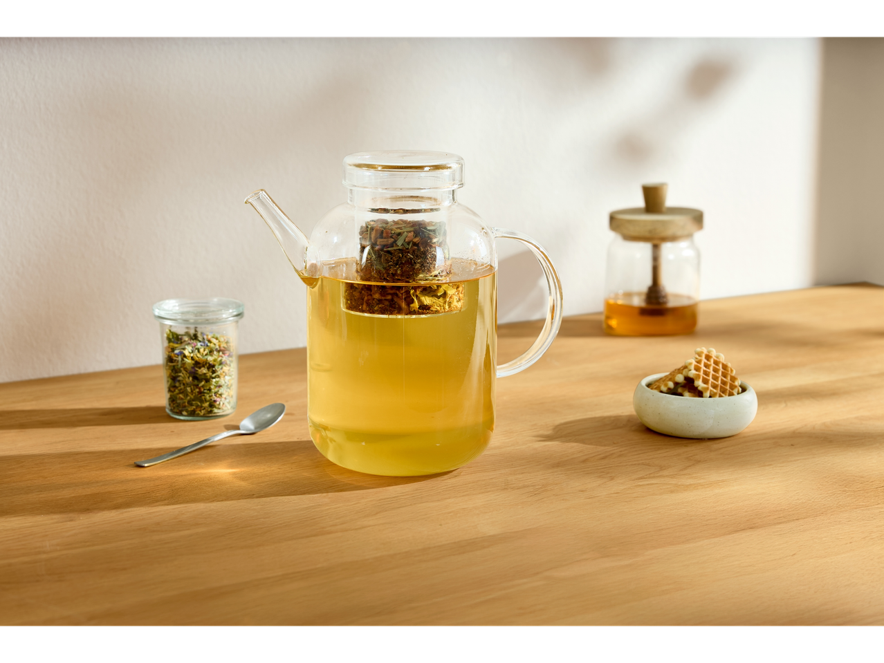 Ceainic de sticlă Silvercrest cu infuzor, miere și vafe pe o masă de lemn.