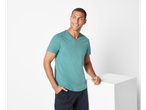 Modelo masculino sorrindo, vestindo camiseta verde e shorts.