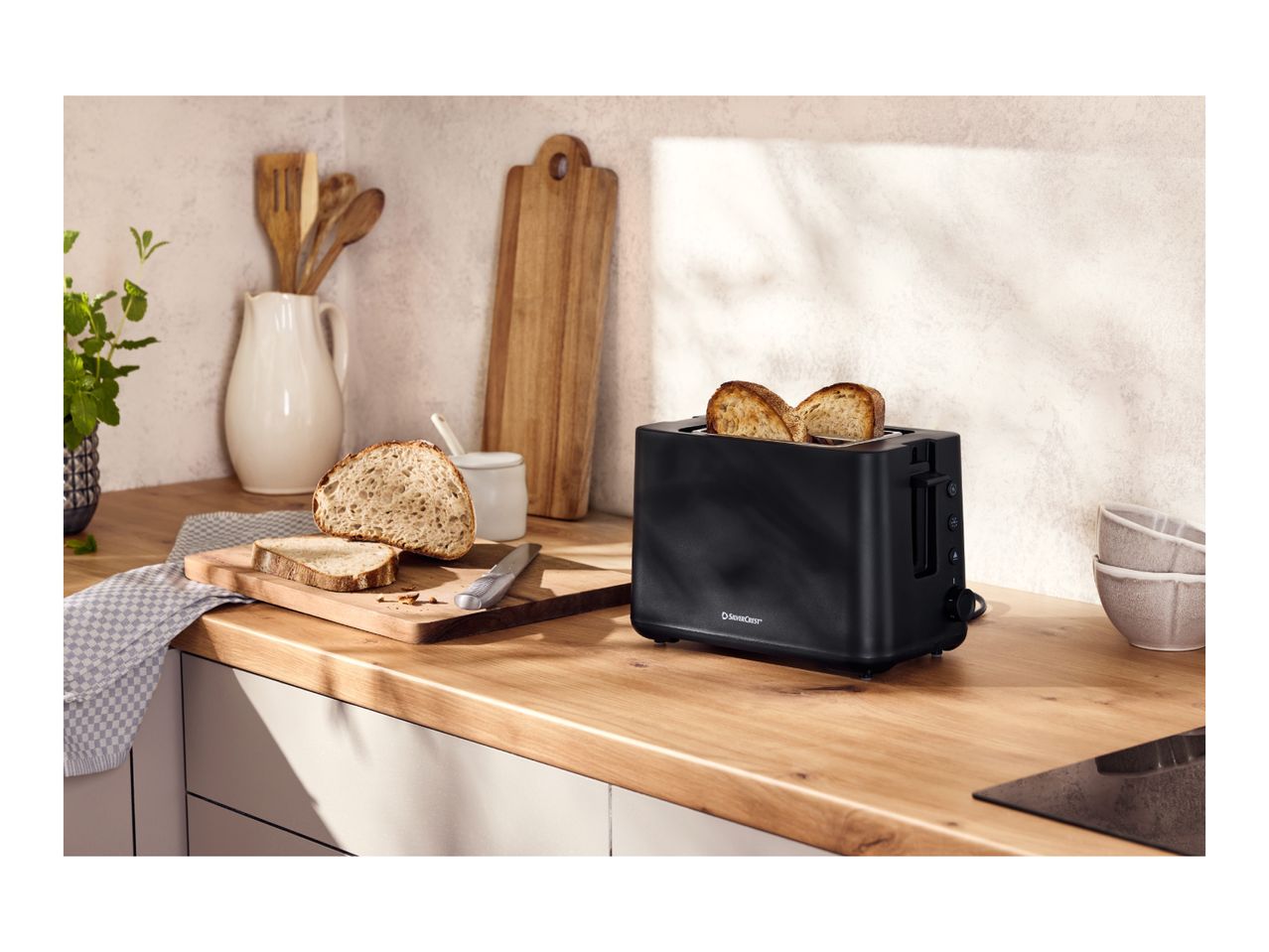 Black toaster with two slices of toast, bread, and kitchen utensils on a wooden counter.