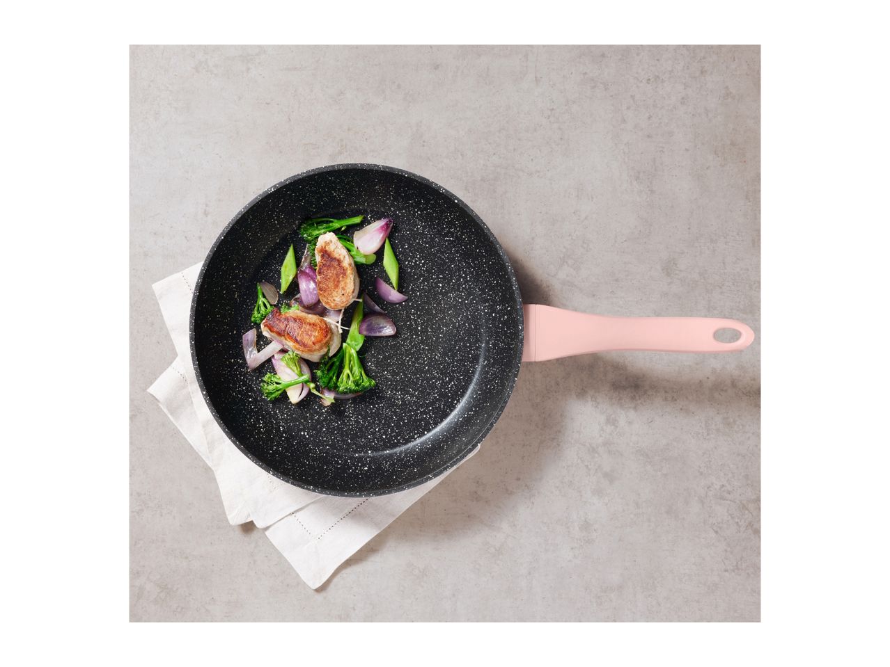 Frying pan with pink handle, containing cooked meat and vegetables on a white cloth.