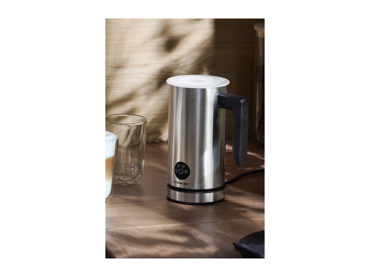 Stainless steel milk frother on a wooden table next to a glass of latte.