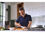 Um homem cortando vegetais em uma cozinha com uma fritadeira de ar e uma panela.
