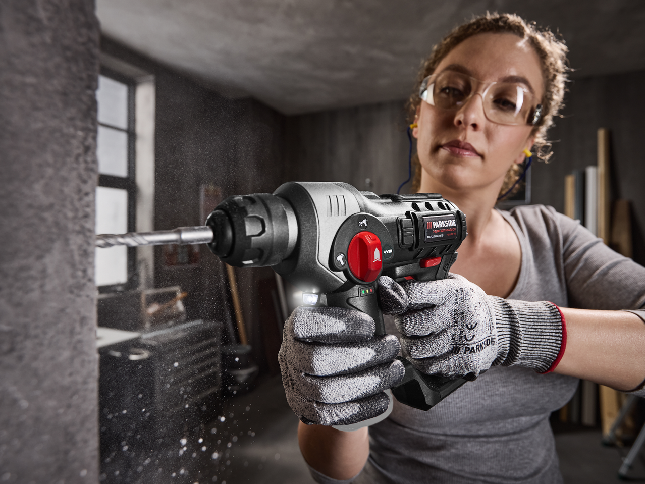 Жена с предпазни очила и тапи за уши пробива стена с акумулаторен перфоратор.