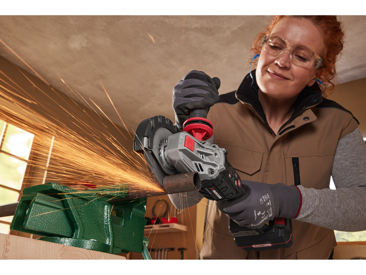 A woman uses a Parkside cordless angle grinder to cut a metal pipe in a workshop.
