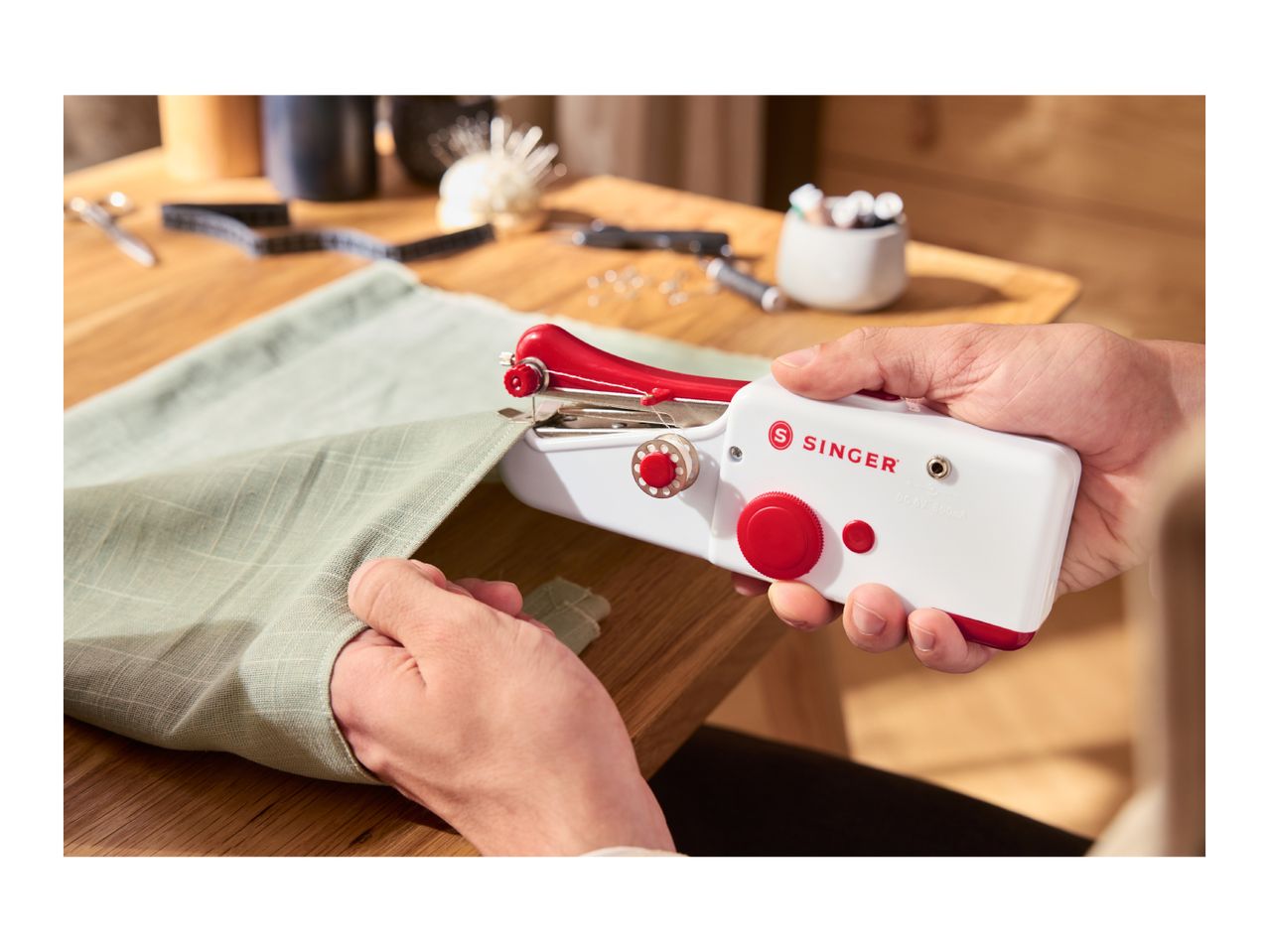 Hands using a red and white Singer handheld sewing machine on light green fabric.