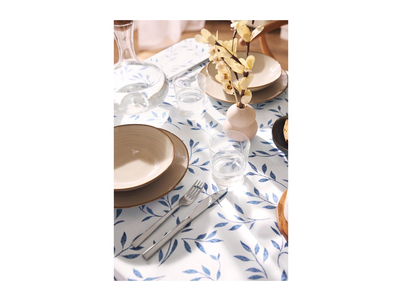Table set with blue leaf-patterned tablecloth, beige plates, glasses, and a vase with flowers.