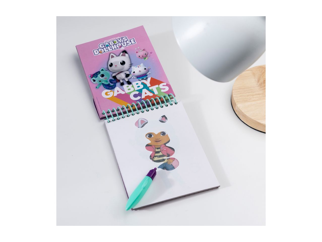 Gabby's Dollhouse coloring book with a pen and a white lamp on a white surface.