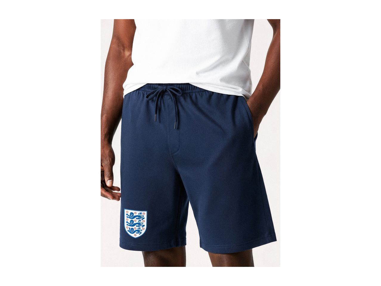 Man wearing navy blue shorts with a white t-shirt, featuring a shield emblem on the left leg.