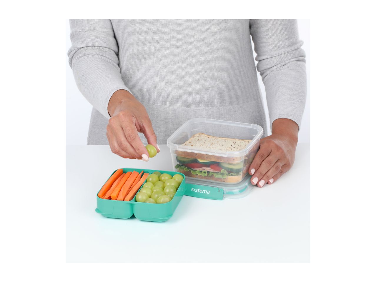 A person packing a sandwich, carrot sticks and grapes into a green lunchbox.