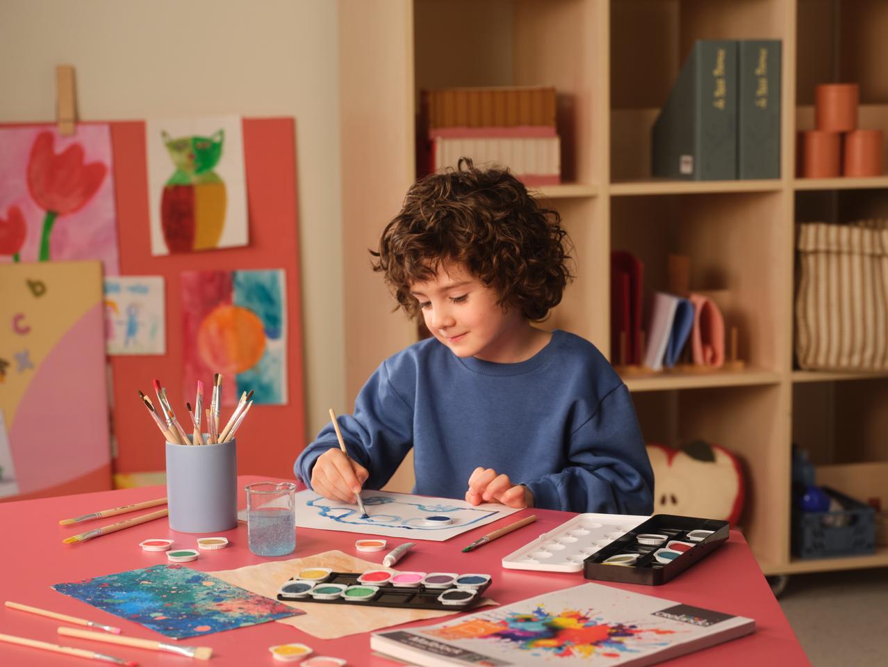 Child painting with Crelando A4 Sketch Pad, watercolors, and brushes on a red table.