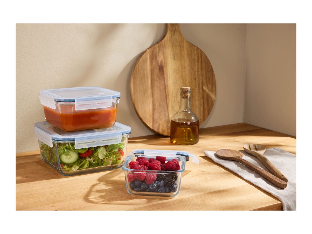 Glass food containers with soup, salad, and berries on a wooden counter with a cutting board.