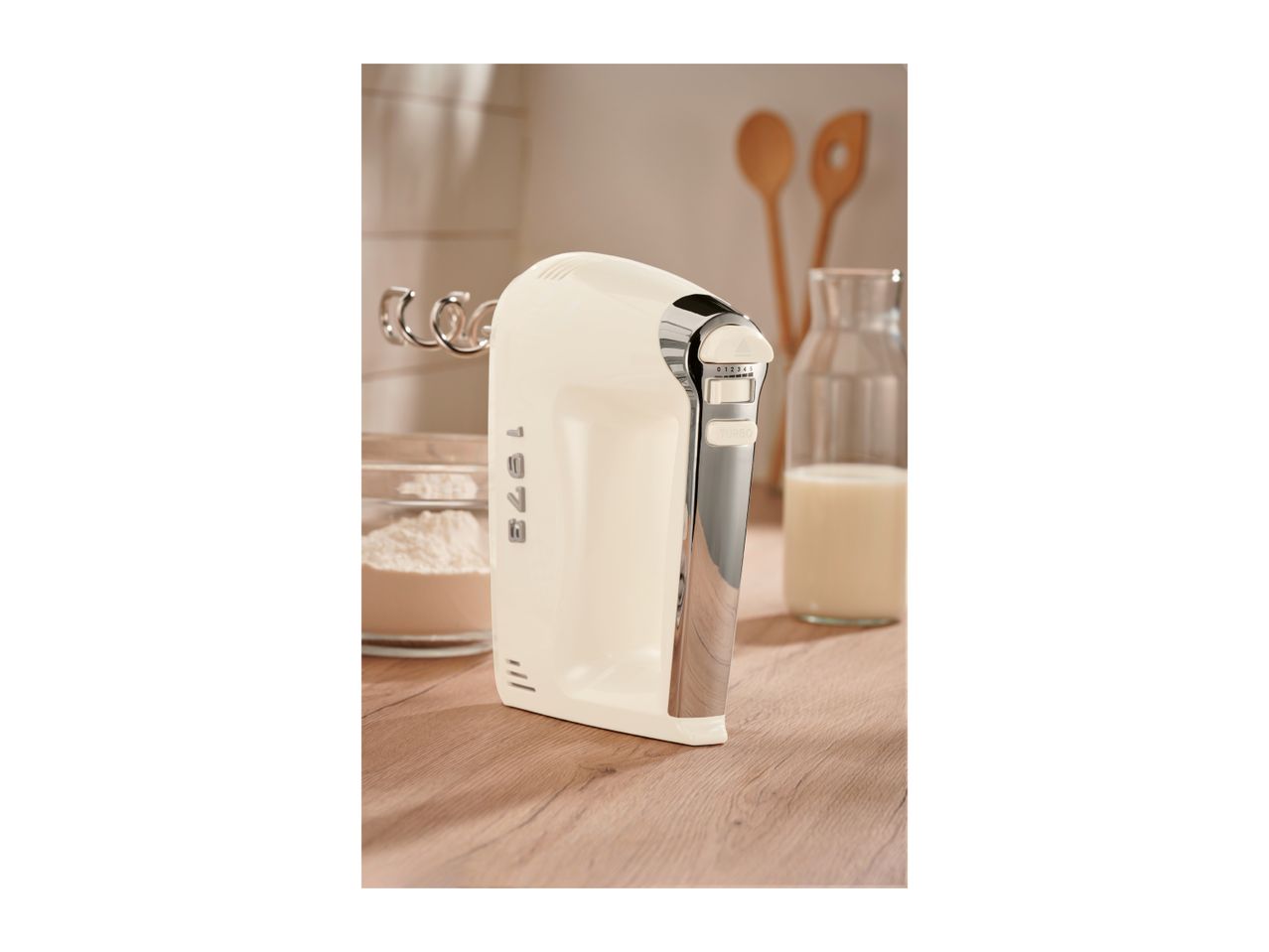 Cream-colored hand mixer with chrome accents on a wooden counter next to flour and milk.