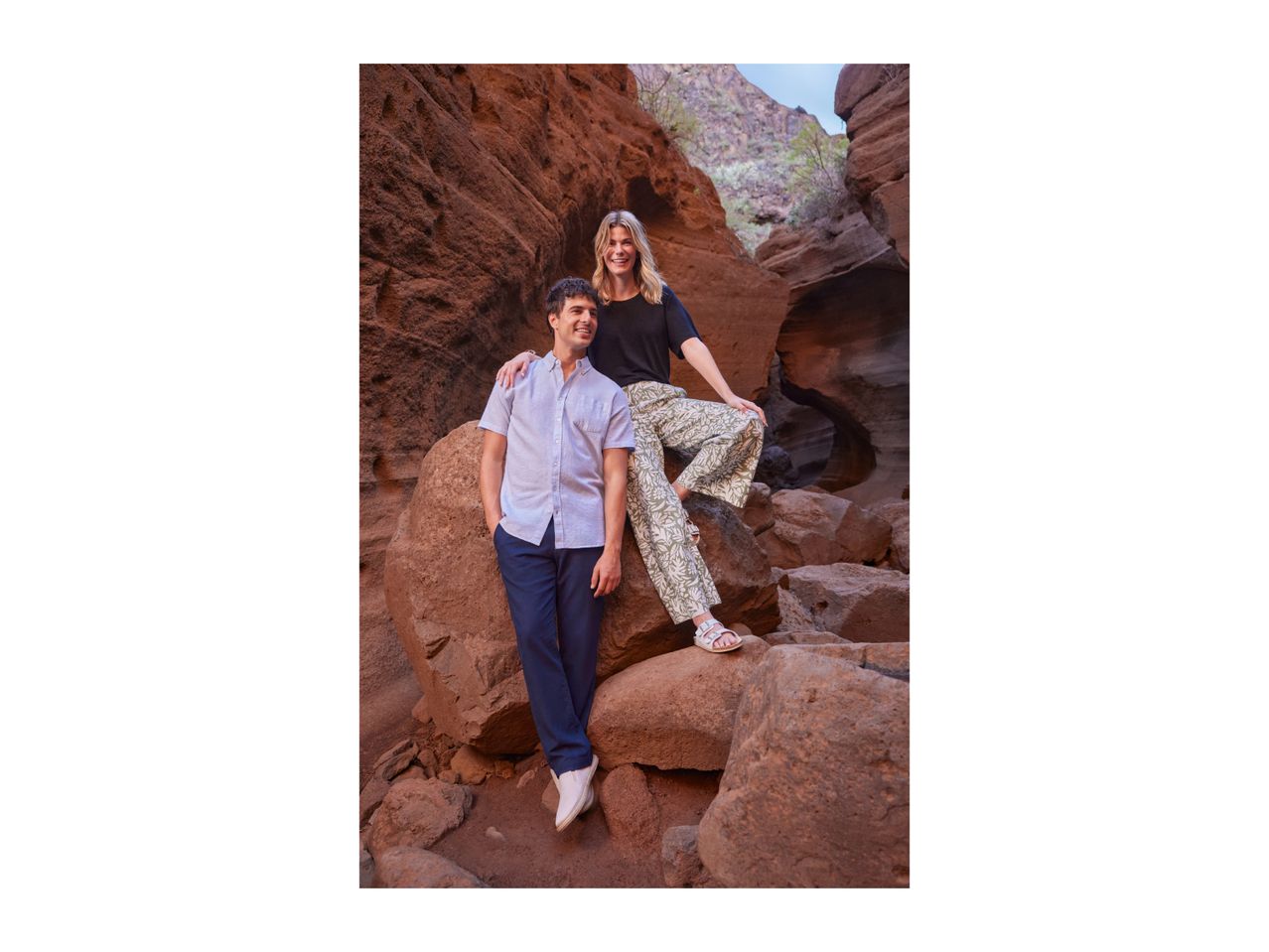 Man in a light blue short-sleeved shirt and navy trousers, woman in a black t-shirt and patterned trousers, posing in a rocky canyon.
