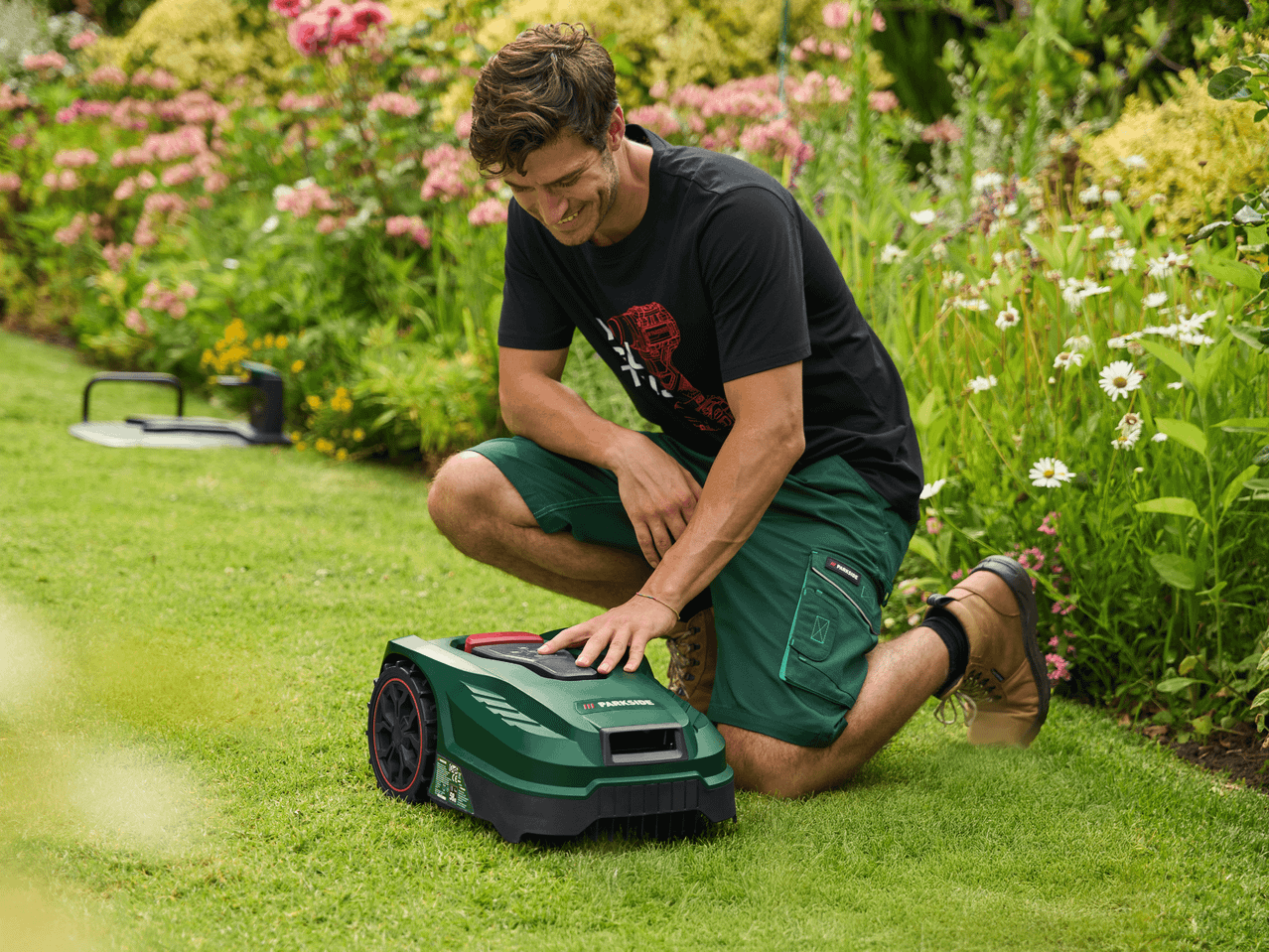 Mann kniet im Garten und bedient einen Rasenroboter auf dem Rasen.