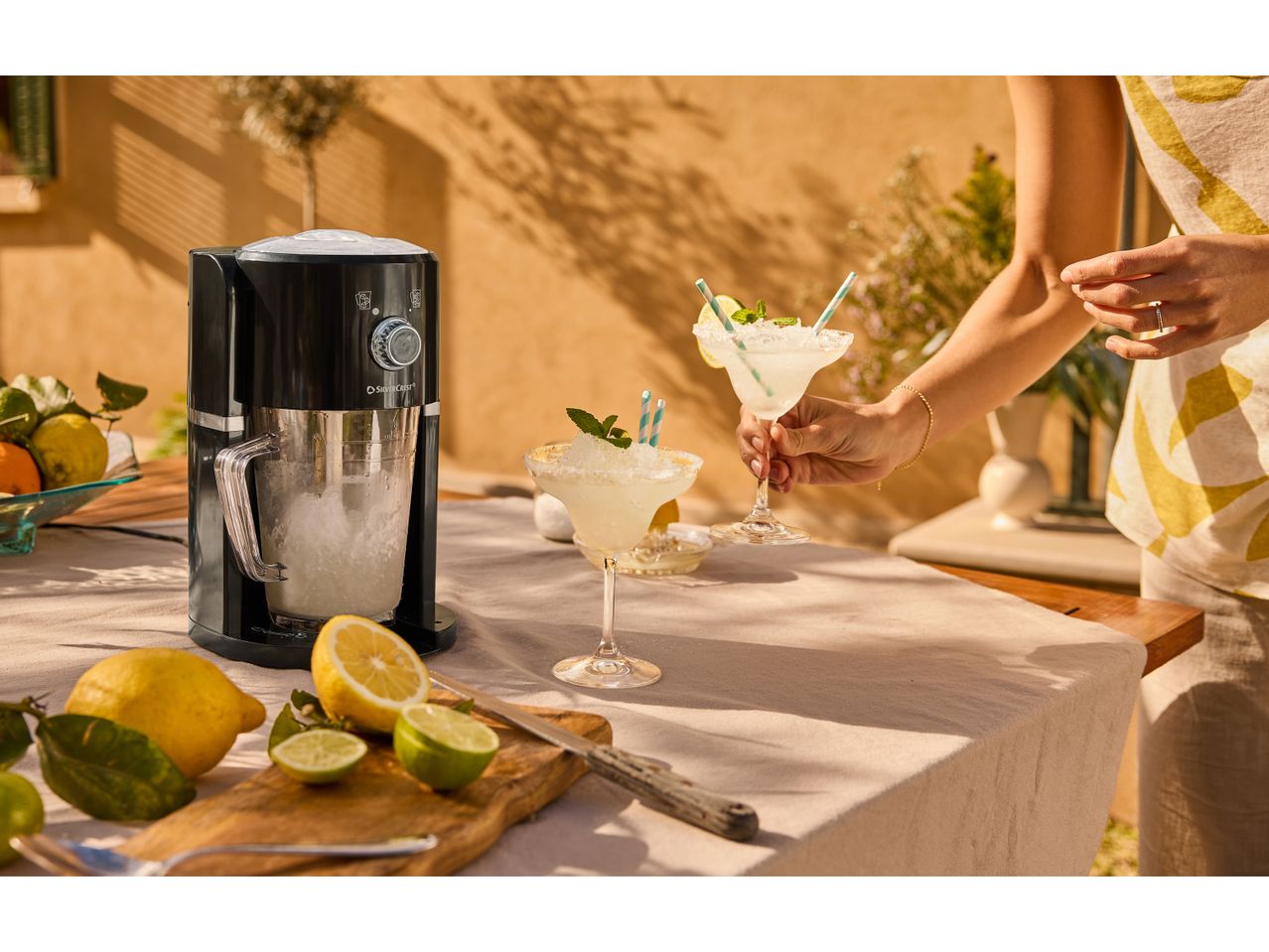 A black cocktail maker sits on a table outdoors, next to fresh fruit and a margarita.