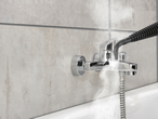 Хромиран смесител за баня с душ слушалка, монтиран на сиви плочки над бяла вана.