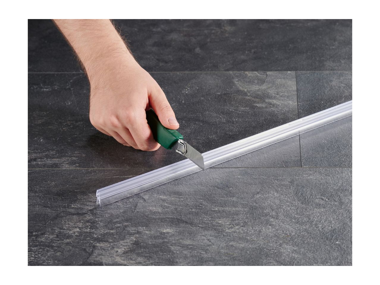 A hand cutting a transparent shower door seal with a utility knife on a dark tiled floor.