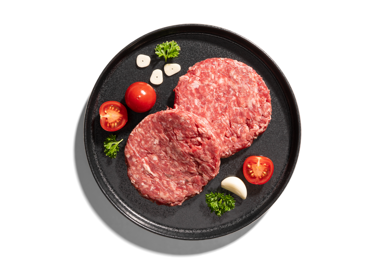 Két nyers hamburgerpogácsa egy sötét tányéron paradicsommal, fokhagymával és petrezselyemmel.