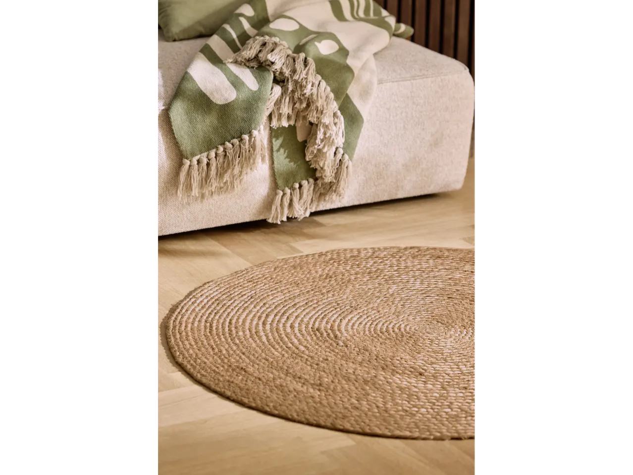 Round jute rug on a wooden floor with a green patterned throw blanket with tassels on a sofa.